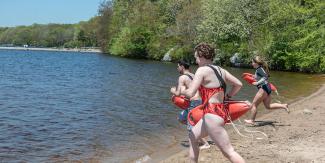 Three lifeguard testing candidates running into Watchaug Pond on a sunny day with a lush, green tree backdrop
