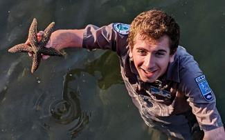 A Park Naturalists looks up at the camera while standing in shallow ocean water holding up a starfish